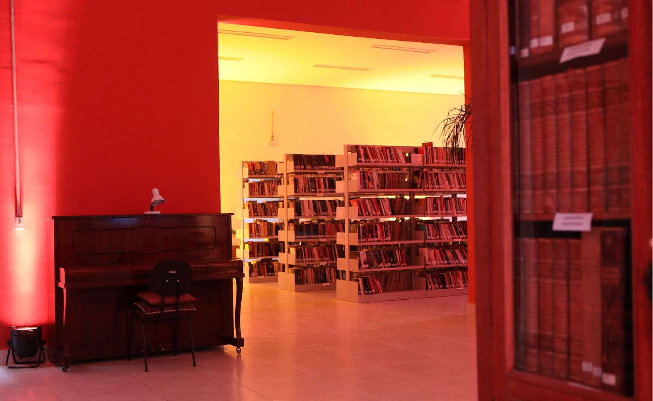 Imagem mostra 3 prateleiras e 1 armário na lateral com livros. No mesmo local também aparece um piano.