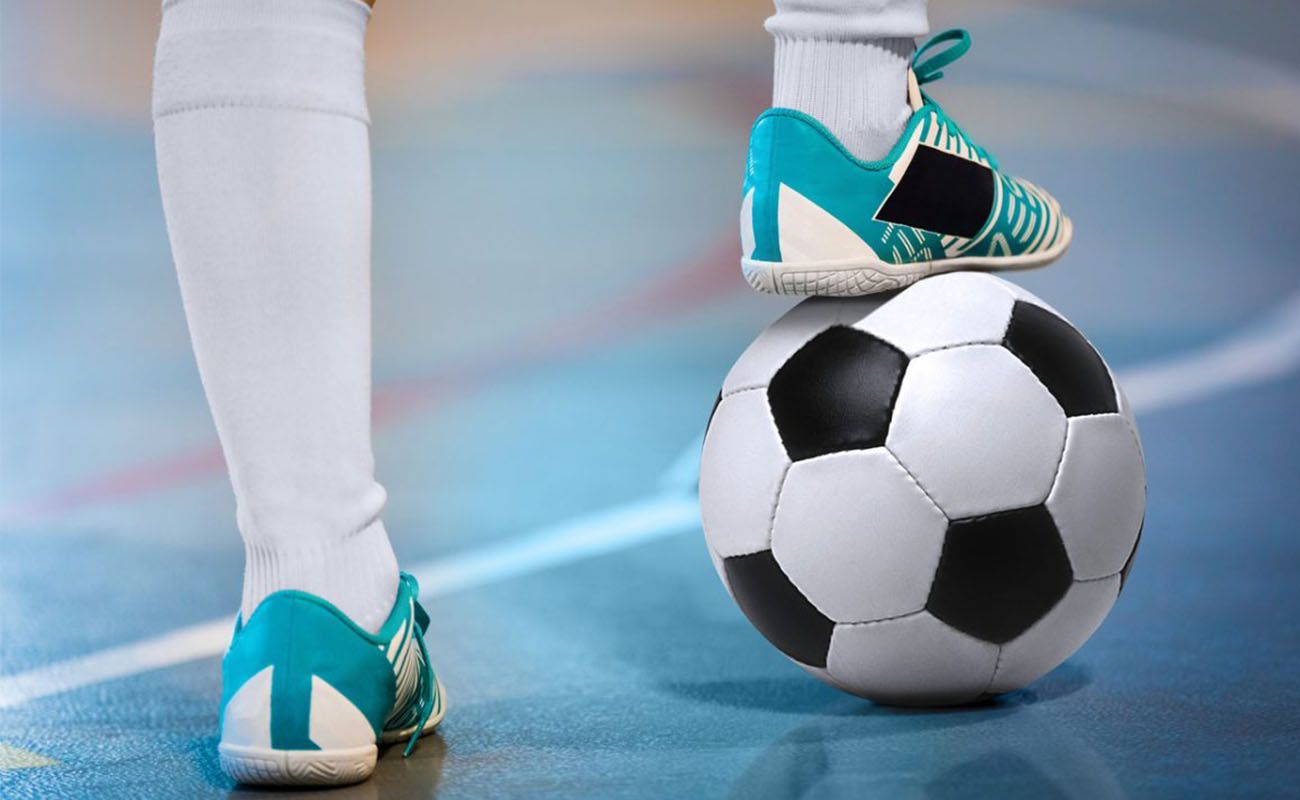 Imagem mostra os pés de uma criança com chuteira de futsal e um deles pisa em uma bola.