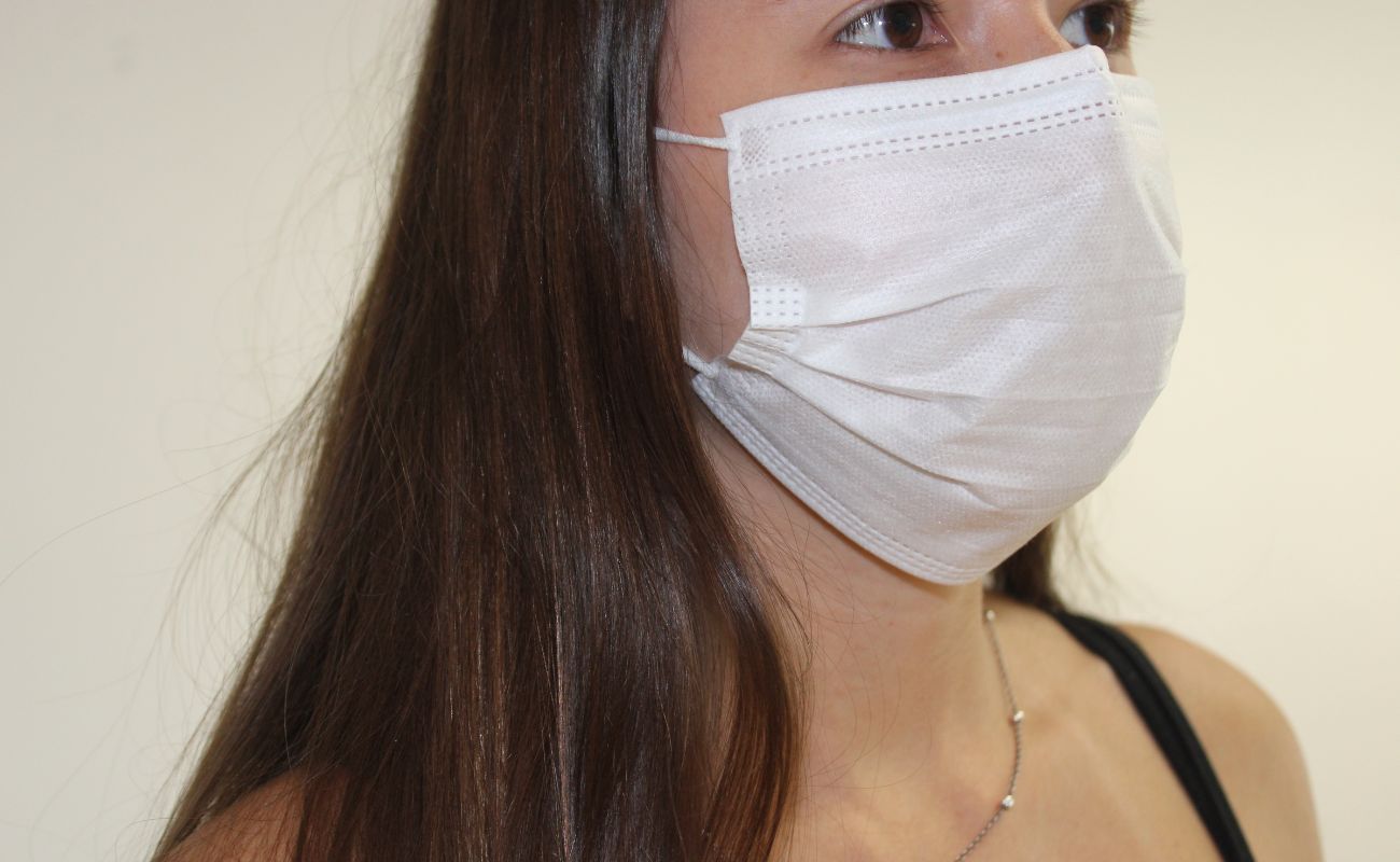 Imagem mostra mulher branca com cabelos castanhos usando máscara de proteção facial no rosto.