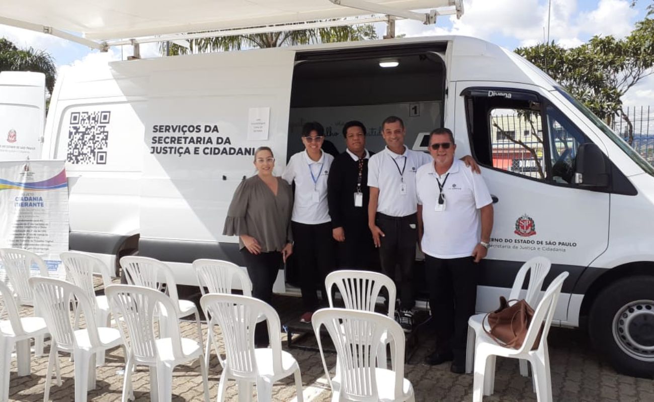Imagem mostra 4 atendentes ao lado da Secretária de Justiça Maria Teresa, e ao fundo a van branca do Projeto Cidadania Itinerante