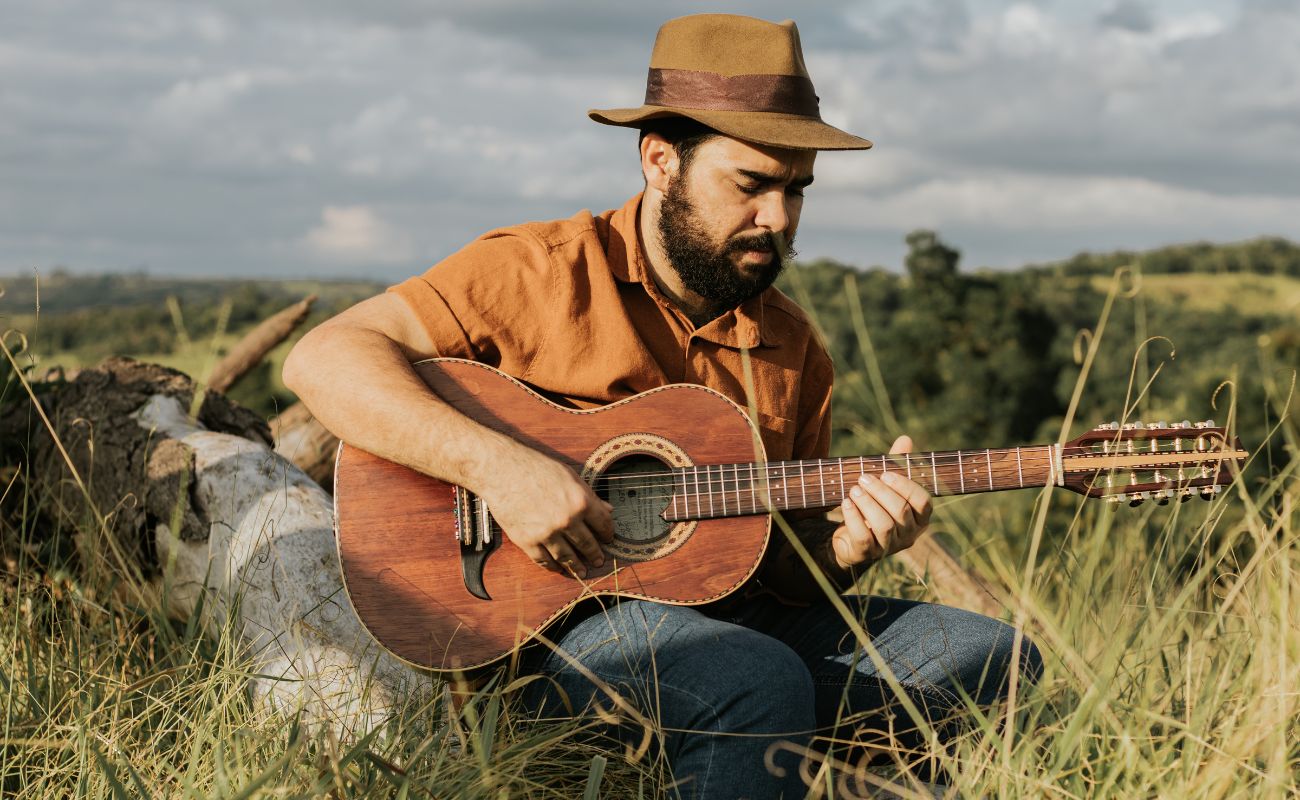 Imagem mostra um homem sentado tocando violão, trata-se do cantor Bruno Addax, que fará apresentação exclusiva no Mercado Municipal, em comemoração aos 3 meses do evento Empório Cultuural