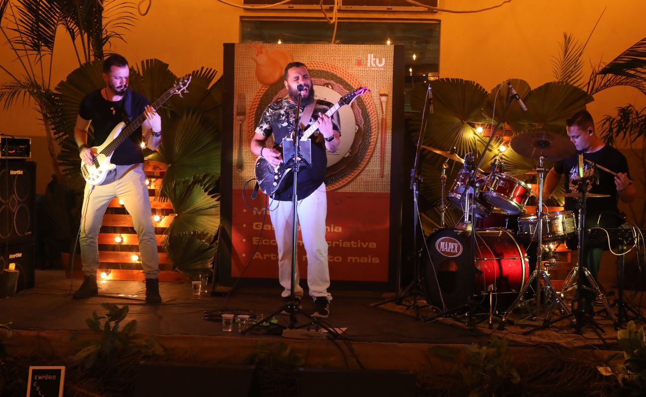 Imagem mostra o palco no dia do evento Empório Cultural, com o cantor Cantor Grilo junto a dois músicos, sendo um deles tocando bateria e outro guitarra.