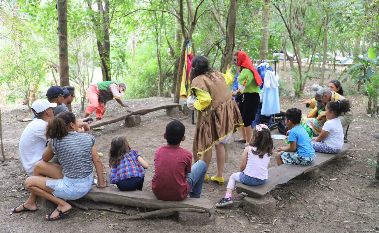 Imagem mostra crianças sentadas de costas, ouvindo histórias contadas por personagens em edição anterior da Feira Ecoart no Parque Taboão.