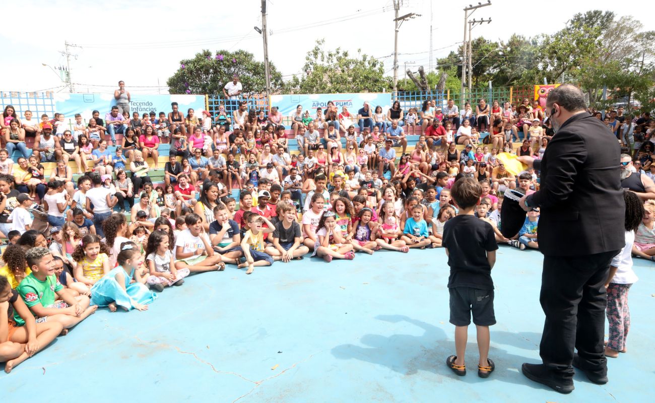 Imagem mostra crianças sentadas observando o show de mágica na Cidade das Crianças em evento anterior comemorando o Dia das Crianças
