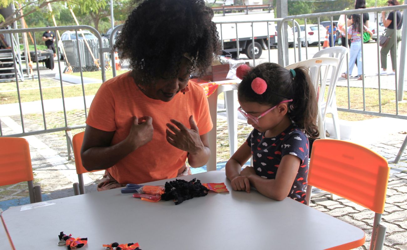 Imagem mostra Rosália Maria Rodrigues de Campos com uma menina em uma mesa confeccionando a Boneca Abayomi que participará da 17ª edição da Primavera dos Museus