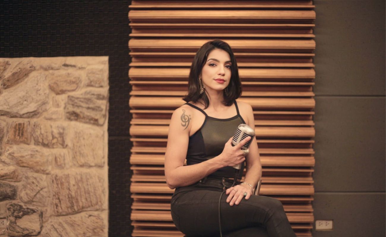 Imagem mostra a cantora Gabi Armenio sentada em um banco segurando um microfone
