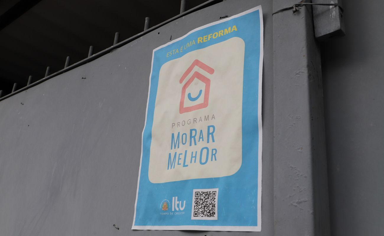 Imagem mostra o portão de uma residência, em foco, com o cartaz indicando que é uma residência que participa do Programa Morar Melhor.
