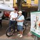 Imagem mostra um agente e saúde passando informações para um munícipe que segura sua bicicleta em praça central da cidade.