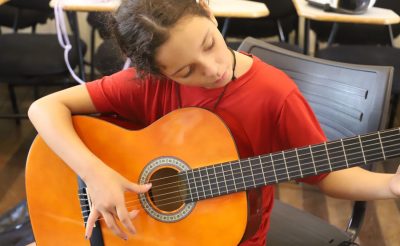 Imagem mostra aluna com camiseta vermelha tocando violão em um dos cursos gratuitos oferecidos pela Emia.