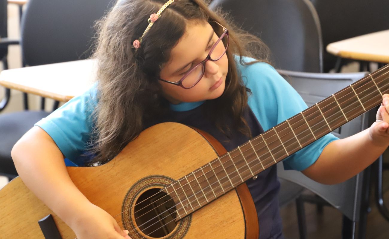 Imagem mostra uma menina de óculos, tocando violão em um curso gratuito da Emia.