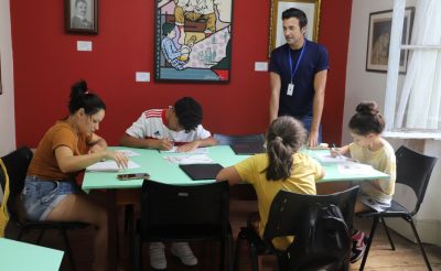 Imagem mostra quatro alunos, dentre crianças e adolescentes, sendo 3 meninas e 1 menino em uma mesa fazendo desenhos sob orientação de seu professor, um homem em pé de camiseta azul marinho, em um dos cursos gratuitos oferecidos pela Emia.