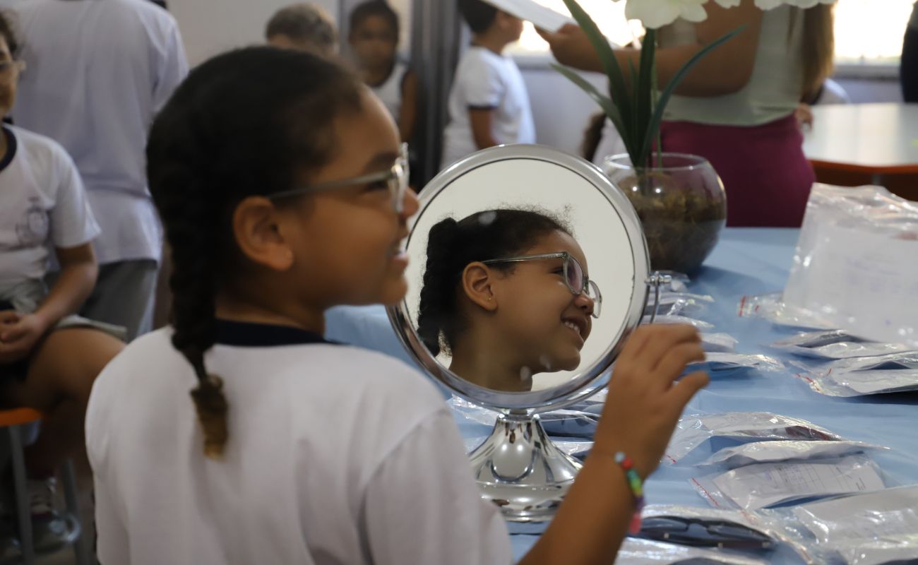 Imagem mostra menina com seu óculo sem frente ao espelho, olhando para a lateral. A referida é beneficiada pelo Projeto Ver + Educa