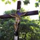 Imagem mostra cruz de madeira e a imagem de Cristo, em meio as copas das árvores, o Cruzeiro do Cemitério Municipal foi objeto do restauro apresentado.