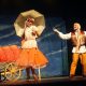Imagem mostra um homem e uma mulher segurando um guarda-chuva atuando em um palco, referente a peça musical.