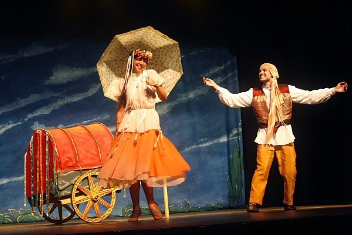 Imagem mostra um homem e uma mulher segurando um guarda-chuva atuando em um palco, referente a peça musical.