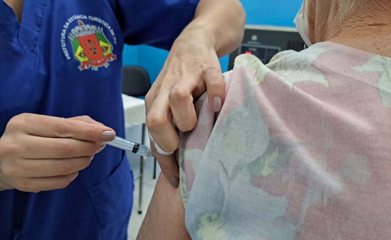 Imagem mostra uma apenas as mãos de uma profissional de saúde aplicando a dose de reforço da vacina contra a Covid em uma mulher de costas.