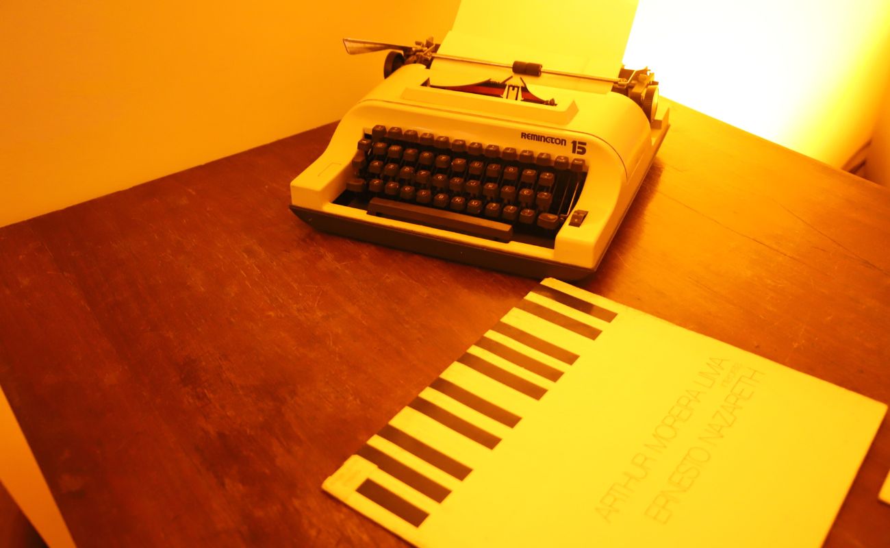 Imagem mostra em close uma máquina de escrever e um livro sobre uma mesa de madeira. No local são realizadas o Clube da Leitura, onde são realizadas uma leitura mensal de um livro e posteriormente os inscritos se reúnem para conversar sobre.