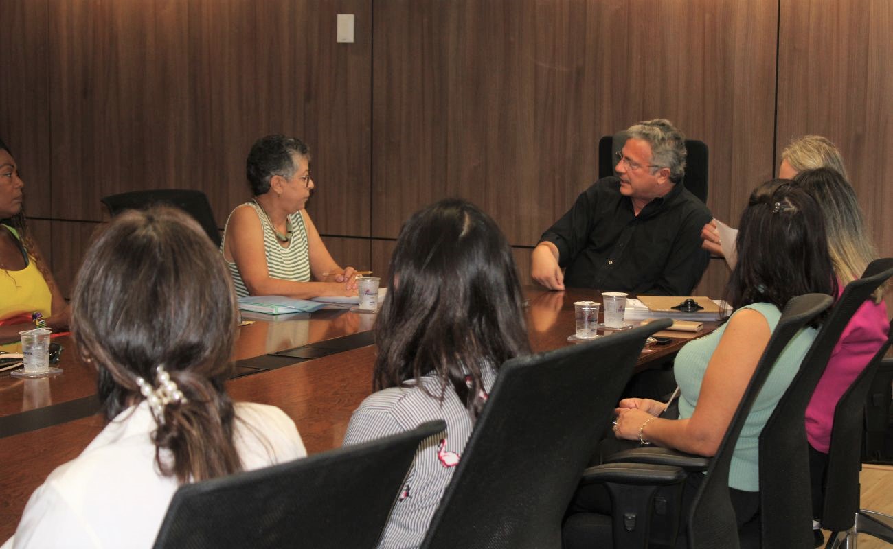 Imagem mostra o prefeito Guilherme Gazzola conversando com a secretária de Promoção Social, junto a 6 mulheres, em uma mesa na sala de reuniões do gabinete. Trata-se da posse de novos membros do Conselho dos Direitos da Mulher