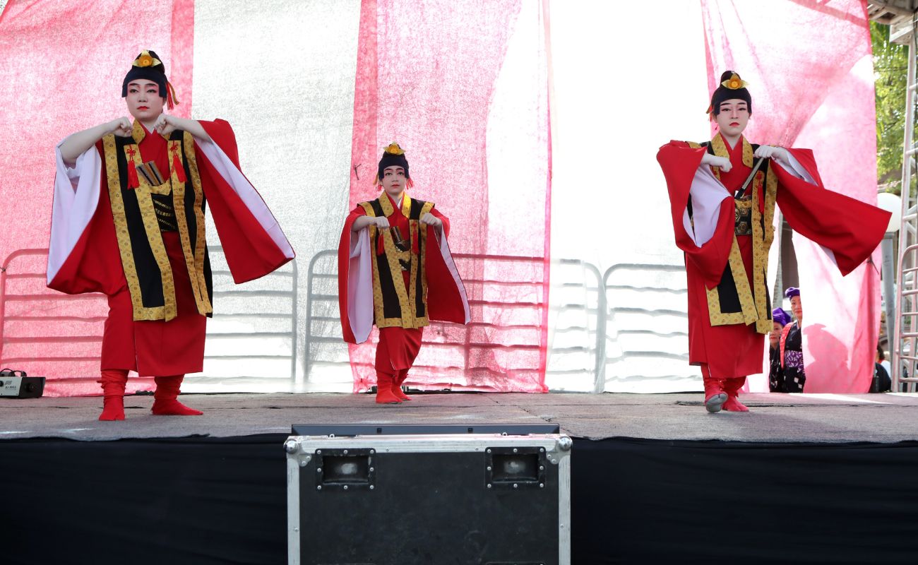 Imagem mostra mulheres japonesas, em uma festa japonesa anterior, vestidas de gueixa, fazendo uma apresentação em um palco.
