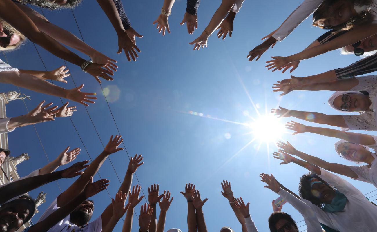 Imagem mostra em ângulo baixo, de várias pessoas com as mãos levantadas, sendo elas em foco, referindo-se ao Dia da Cultura e da Paz.