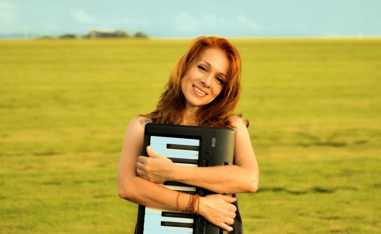 Imagem mostra a cantora Olivia Genesi segurando seu teclado em um gramado.