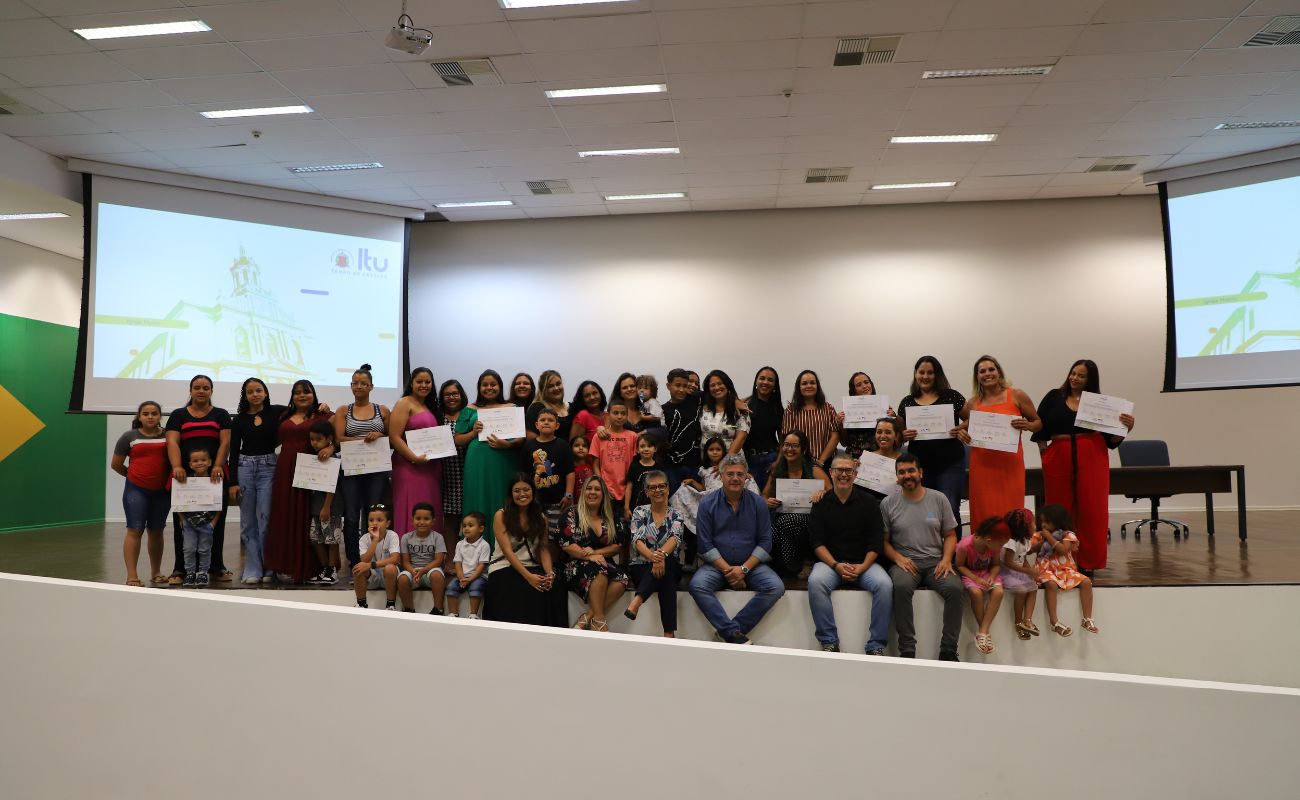 Imagem mostra o prefeito Guilherme Gazzola no palco do auditório do Paço Municipal, com sua equipe e com mulheres participantes do Programa Prospera Família.