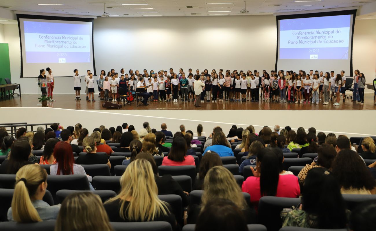 Imagem mostra profissionais da educação de costas sentadas no auditório do Paço, participando da Conferência do Plano Municipal
