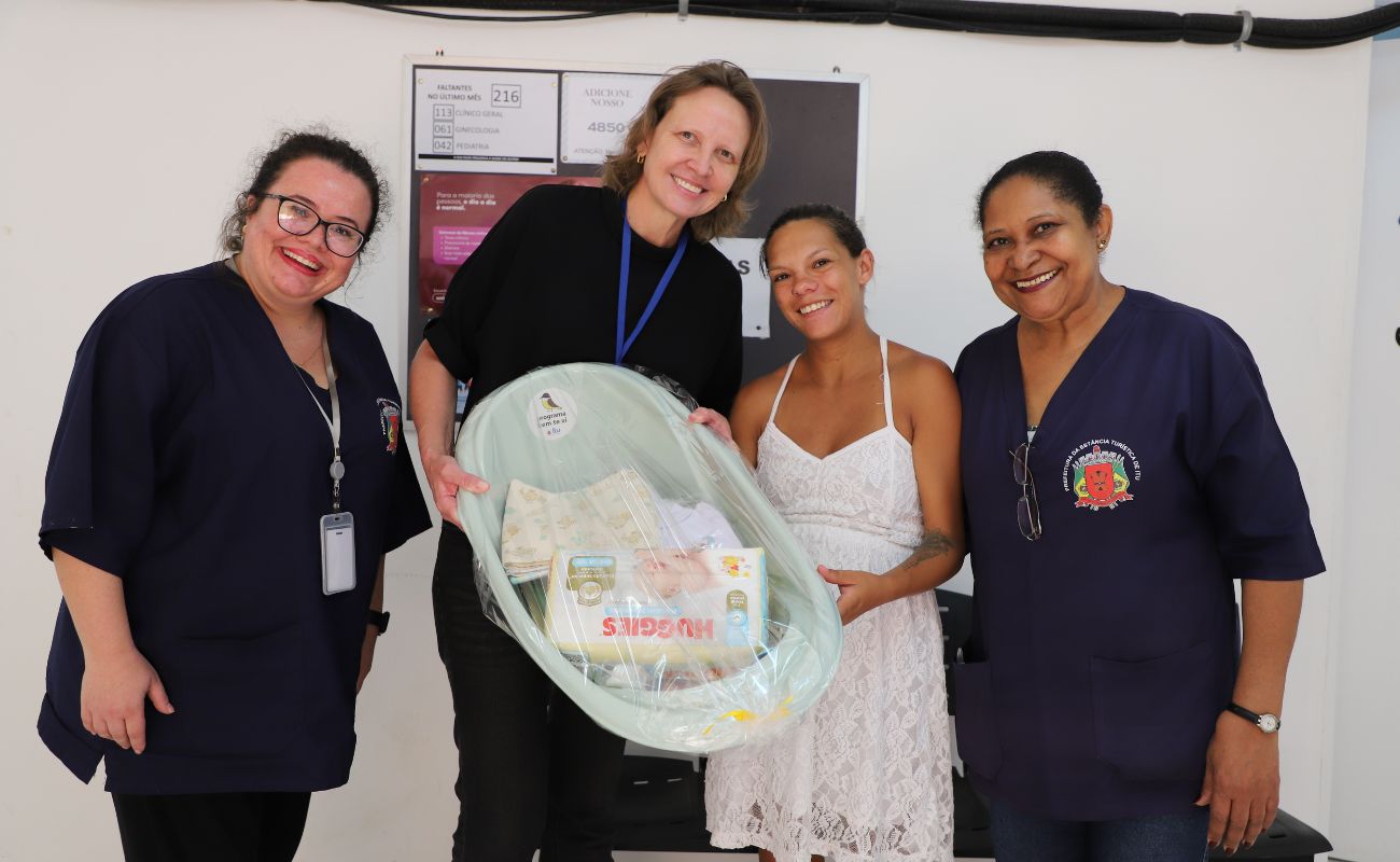 Imagem mostra a secretária de saúde Janaina junto a duas enfermeiras, entregando um kit a uma mãe, referente ao Programa Bem-Te-Vi