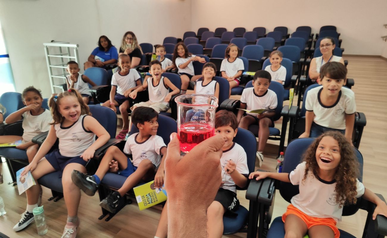 Imagem mostra crianças olhando curiosamente para um copo com líquido vermelho, referente a experimento realizado em uma das ações de promoção do consumo consciente e da preservação ambiental em Itu, durante a Semana da Água em Itu.