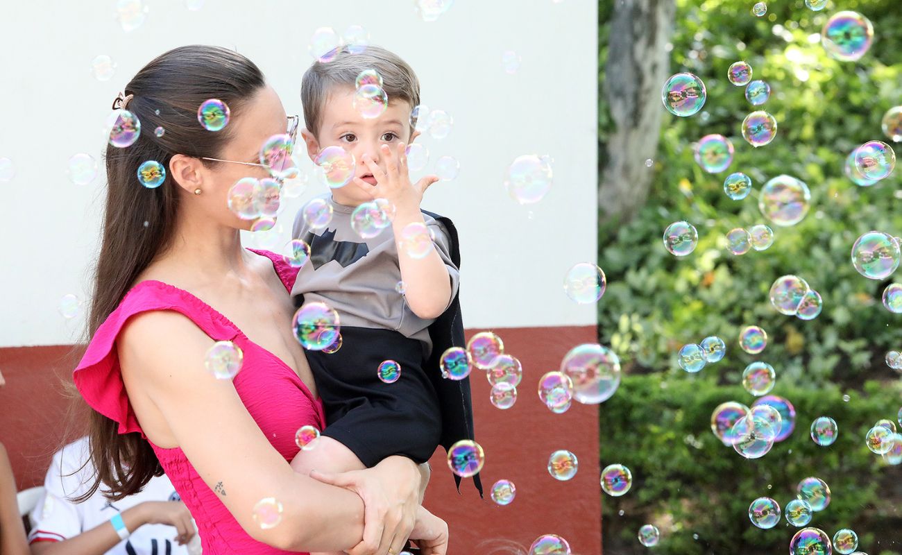Imagem mostra uma mãe com uma criança no colo, observando bolhas de sabão em evento de Conscientização do Autismo.