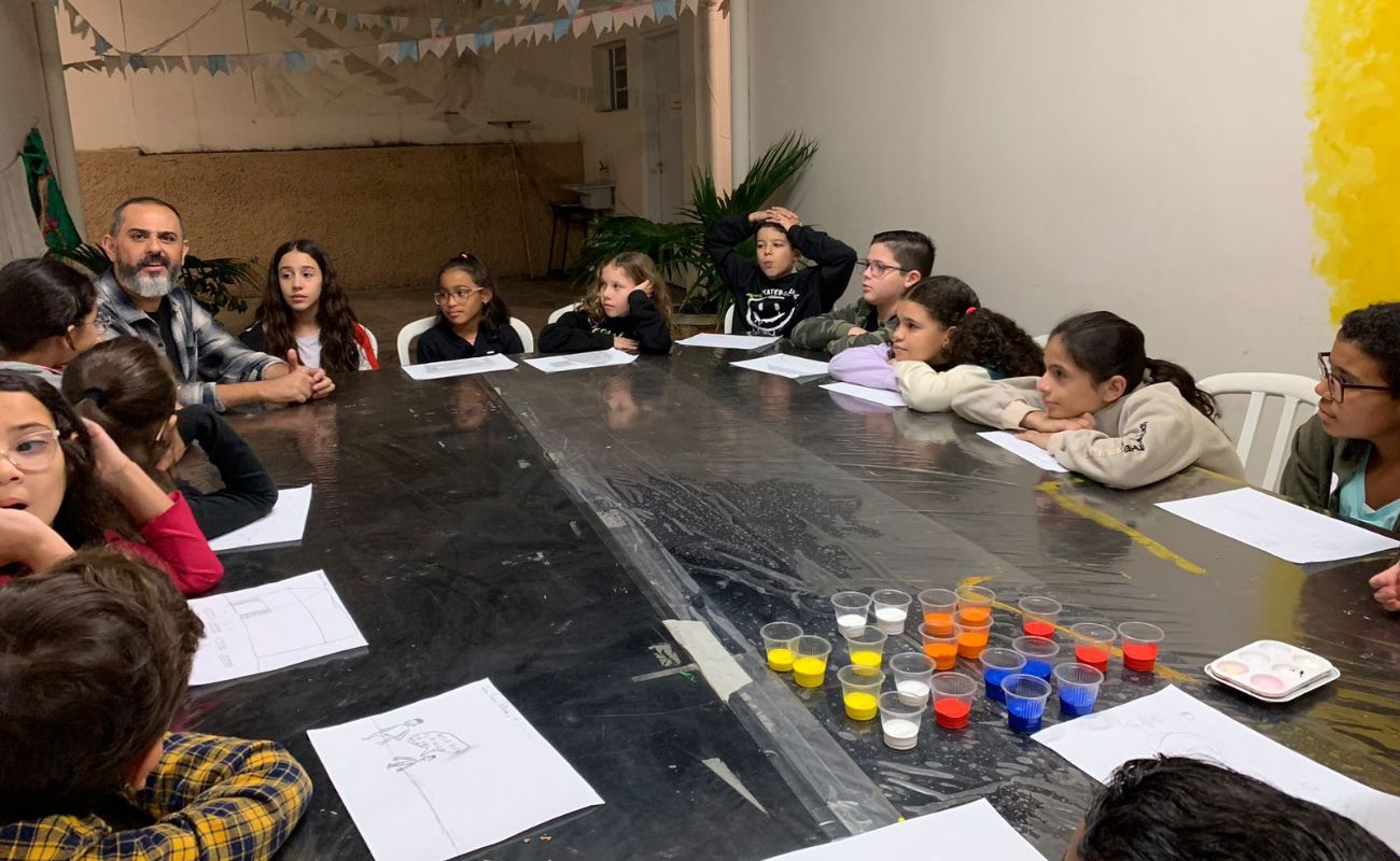 Imagem mostra o coordenador Thomas Rodrigo junto das crianças sentadas aguardando o momento da sua atividade, no segundo encontro do Comitê das Crianças.