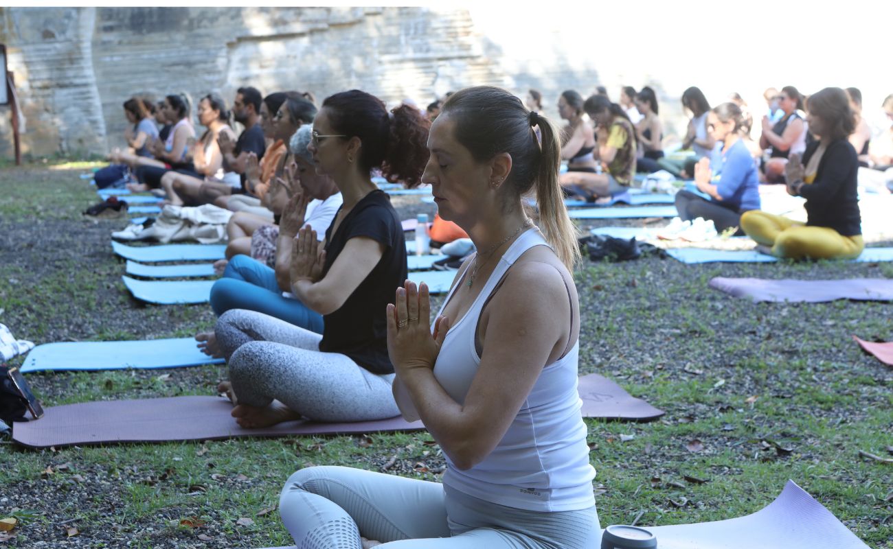 Imagem mostra mulheres em posição de yoga, com as mãos juntas e olhos fechados.