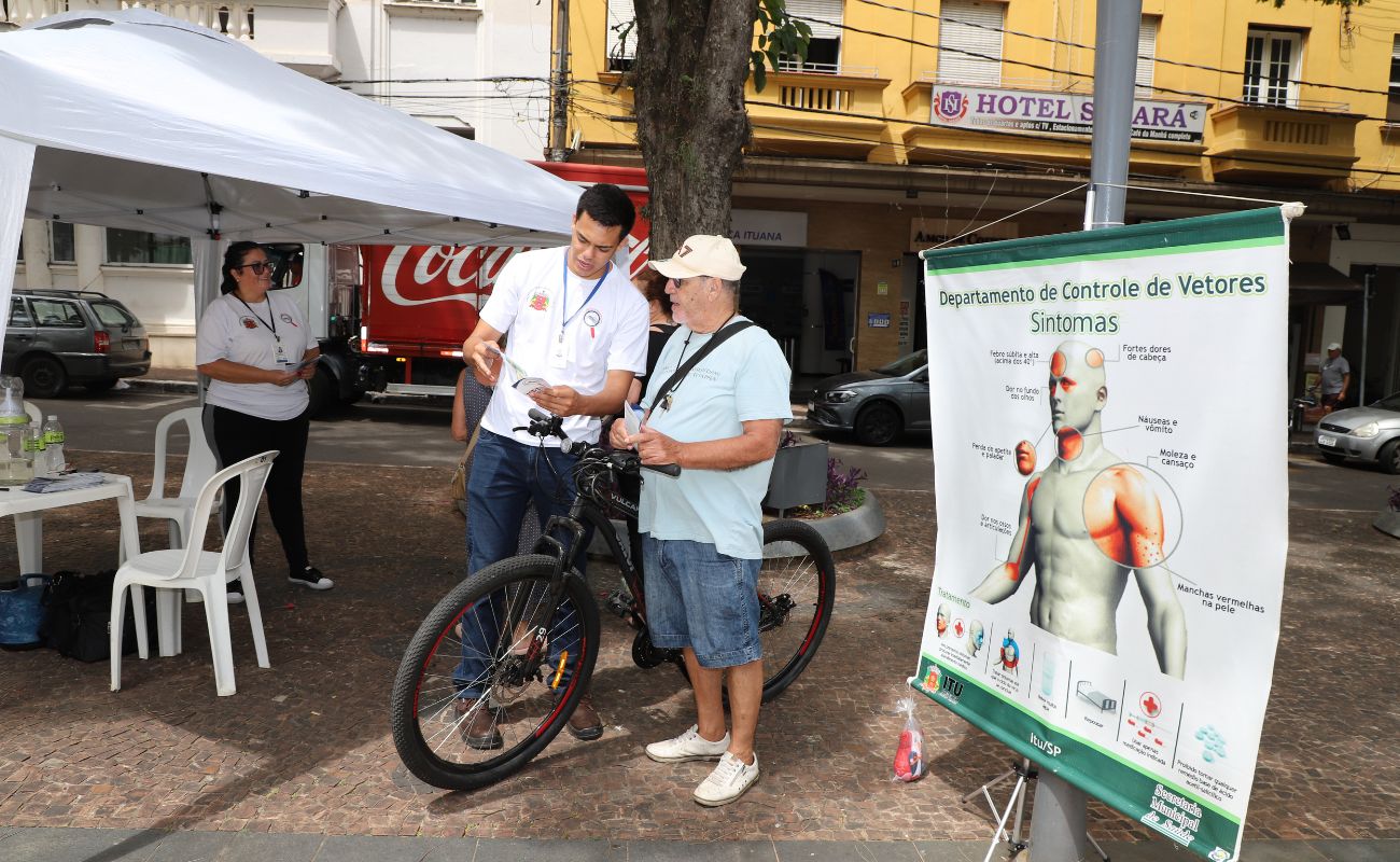 Imagem mostra um agente e saúde passando informações para um munícipe que segura sua bicicleta em praça central da cidade.